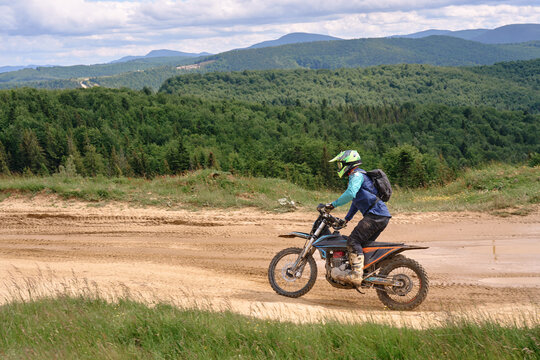 A dirt bike rider with helmet and backpack rides a sandy off-road trail past lush green forests and distant mountains, capturing action, adventure and freedom in an outdoor motorsport scene.