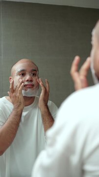 Vertical Man applying a sheet mask in front of a bathroom mirror during his morning skincare routine. Healthy aging longevity. Men self-care anti-aging skincare. wellness and preventive health.
