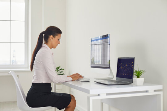 Accountant analyst businesswoman reviews spreadsheets and graphs. She works at a monitor and laptop carefully checking data records in a clean office. Data driven finance analysis and audit.