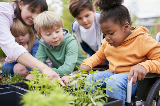 Group of children, including a girl in a wheelchair, engaged in gardening activity, planting herbs and vegetables in black containers outdoors in a green environment