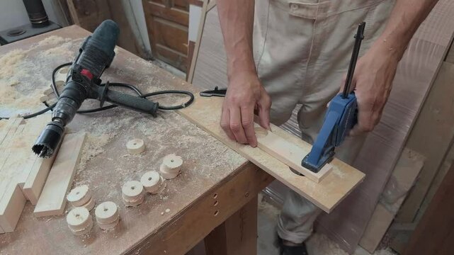 Dedicated self employed entrepreneur in workwear drilling circular shapes from clamped plywood board with heavy power tool inside local home manufacturing studio during shaky handheld close up