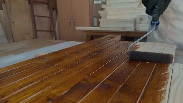 Detail of hands in black gloves applying rich dark brown wood stain to raw pine timber planks with small roller during commercial furniture finishing in private home production workshop studio