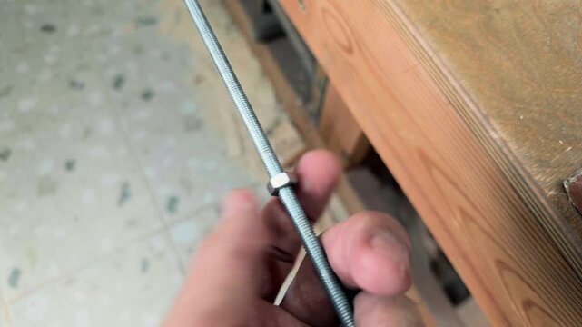 Extreme close up of an unrecognizable independent creator manually spinning a small hexagonal steel nut onto a long threaded metal stud during DIY hardware assembly inside a private craft room