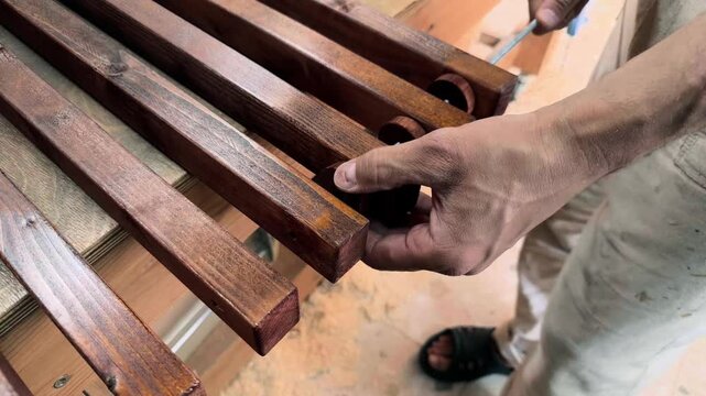 Handheld high angle shot of an anonymous self-employed maker threading dark stained wooden slats and spacers onto a metal rod during authentic DIY furniture assembly inside a domestic workshop