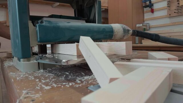 Professional woodworker wearing overalls feeding raw wooden planks into a thickness planer machine on a workbench, surfacing timber blanks in an authentic carpentry workshop, close up tracking shot.