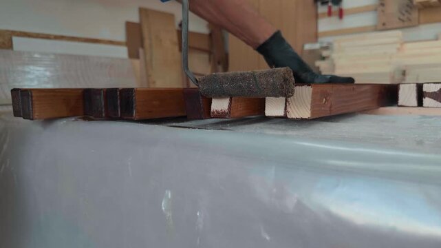 Skilled maker in protective gloves applying dark wood stain to raw pine timber edges with handheld roller in professional woodworking workshop for custom furniture manufacturing project