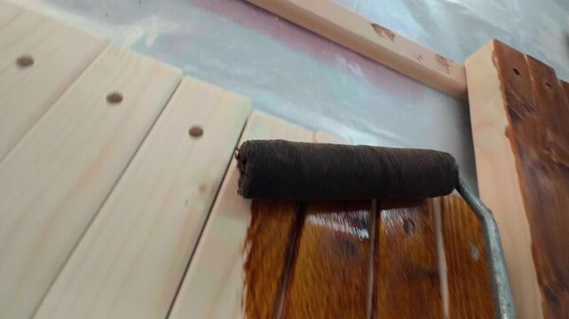 Unique tool mounted camera point of view showing paint roller applying dark brown wood stain to light pine timber boards during commercial furniture finishing in local home production workshop
