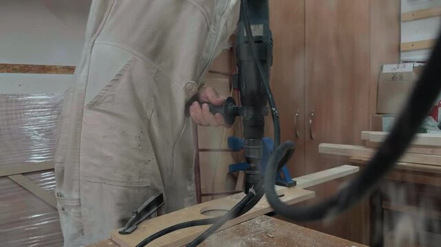 Unrecognizable carpenter in coveralls using heavy duty power drill with hole saw bit to cut circular shapes in plywood board in dusty woodworking shop with hanging cable and sawdust transition
