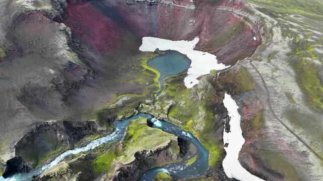 Aerial view of the vibrant red volcanic crater of Raudibotn with a winding river, mossy green terrain, and patches of snow in Svartafellstangi, Skaftarhreppur, Iceland.