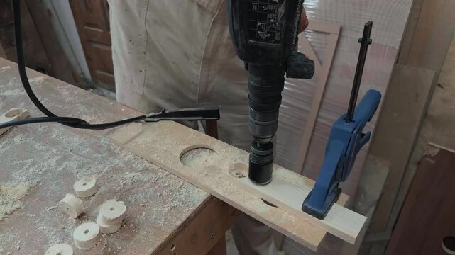 Unrecognizable woodworker in protective coveralls cutting circular wooden discs from a clamped plywood board using a heavy rotary drill on dusty workbench during shaky handheld close up shot