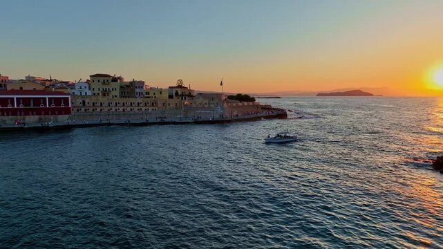 Aerial view of Old Venetian Port of Chania at sunset with a small boat on the water and historic buildings along the coast in Chania, Greece.