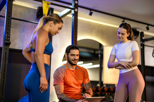 A seated personal trainer explaining a workout schedule on paper to two standing women in a gym.