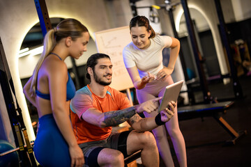 Fototapeta premium A seated personal trainer showing a digital workout plan on a tablet to two standing women in a gym.