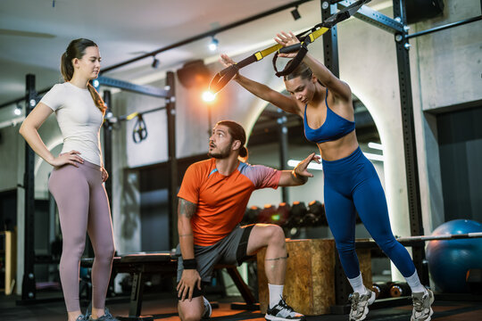 A personal trainer pointing at a woman core while another client exercises with suspension straps in a gym.