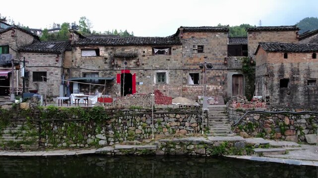 A row of weathered traditional stone buildings stands by a waterway in a rural ancient village, with stone stairs leading up to the structures.