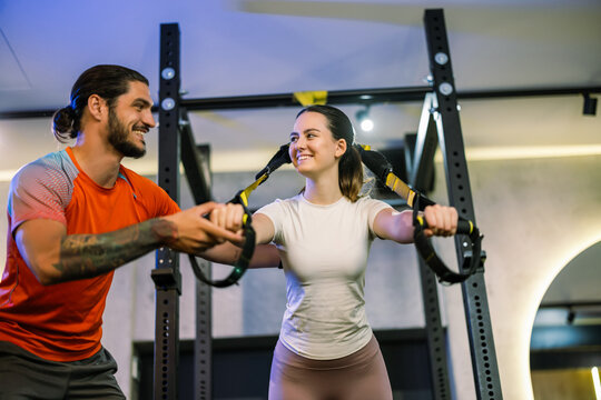 Standing fitness instructor guiding a woman during a suspension strap workout in a gym