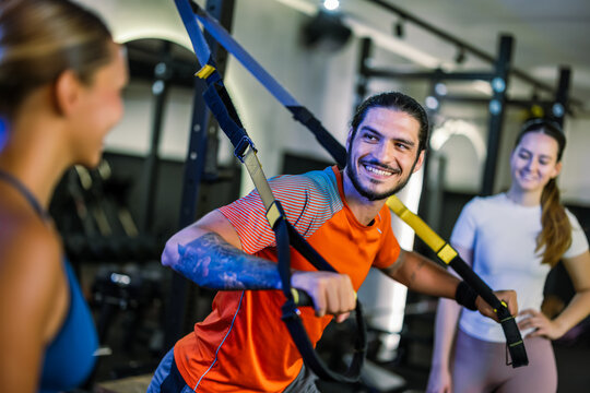 Two young women listening to a male trainer explaining a suspension strap exercise in a gym