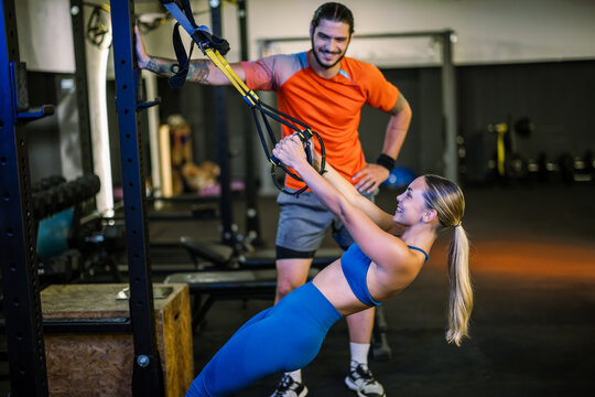 Standing fitness instructor supervising a woman performing a suspension training back exercise