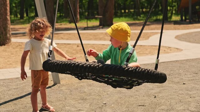 Two children enjoy playing at a park on a sunny day. One child pushes the other who is sitting on a large swing. They are having fun in the outdoor space