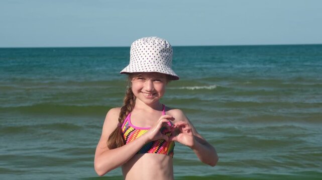 Happy girl in panama hat making heart gesture with hands on sea background. Concept of love for summer travel and holidays