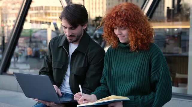 Young man and woman studying together using a laptop and taking notes on a notepad outdoors