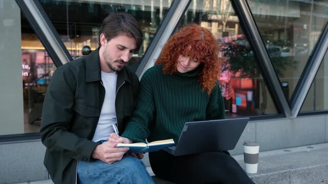 Young man and woman working together, studying and using a laptop on a city street