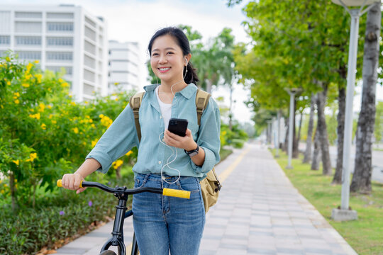 Asian woman wearing headphones, holding a phone, smiling at a funny video on a bicycle in a park.
