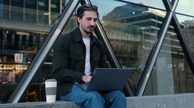 Confident man sitting in front of a modern building typing on a laptop and drinking coffee
