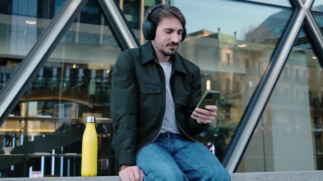 Young man sitting in front of a modern building using his smartphone and listening to music