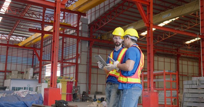 Two industrial workers collaborating on site management, inspecting project details on a digital tablet inside a large steel fabrication factory.