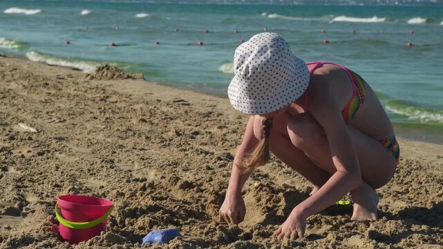 Girl in panama hat playing with sand near bucket on sea beach. Concept of creative children holiday activities and summer vacation