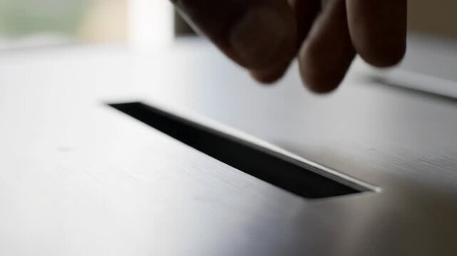 Hand inserts textured paper into a slot, close-up, neutral background, concept of submission or deposit.