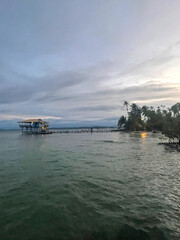 Fototapeta premium Palm trees on a sandy beach in Bocas del Toro, Panama at sunset. Wooden pier and stilt building, ocean waves, tropical sky.
