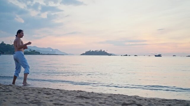 A man practices martial arts and works on his kicking techniques. Sandy tropical shoreline with raised knee, bent arms, calm water, distant islands, small boat, and soft pastel sky at glowing sunset