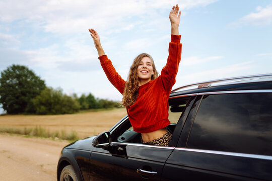Female traveler leans out car window and feels freedom of sunset. Young woman on a road trip enjoys rural scenery outdoors. Concepts of adventure, vacation, and transportation.