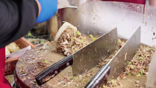 A chef wearing blue gloves prepares food, chopping ingredients with large cleavers on a wooden cutting board in a food preparation setting.