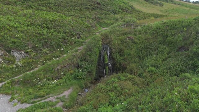 Aerial view of the small waterfall at Lansallos Beach flowing through a lush green valley with ferns and grassy hills in Looe, Cornwall, United Kingdom.