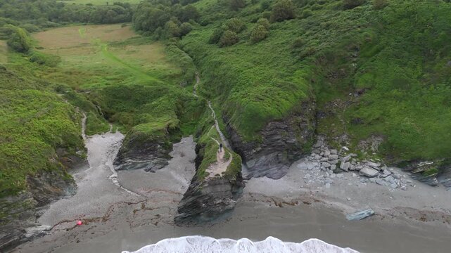 Aerial view of Lansallos Beach featuring rugged cliffs, lush green hills, and waves crashing onto the sandy shore in Looe, Cornwall, United Kingdom.
