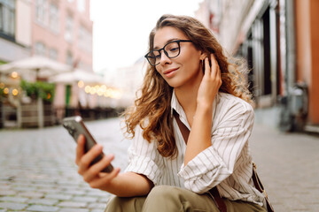 A young woman stands in a city square, holding a phone. A woman in glasses enjoys a sunny day in the city, holding a smartphone. Concept: technology, urban, lifestyle. © maxbelchenko
