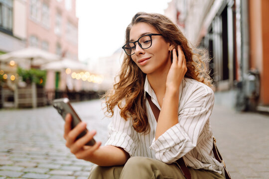 A young woman stands in a city square, holding a phone. A woman in glasses enjoys a sunny day in the city, holding a smartphone. Concept: technology, urban, lifestyle.