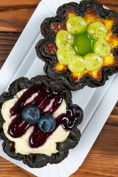 Two flower-shaped black crust egg tarts topped with blueberry cream sauce and green grape fruit glaze on white paper tray