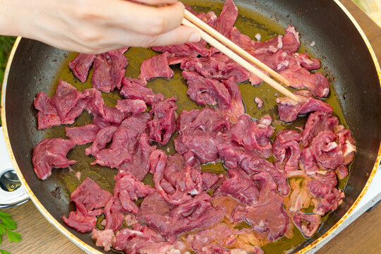 Thin raw beef slices spread in an oiled frying pan, a hand holds wooden chopsticks to stir the fresh meat for stir-frying