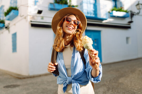 Smiling woman eats ice cream while strolling through old streets of town. Tourist in hat admires city's architecture, eats ice cream at sunset. Concept of relaxation, tourism, and enjoyment.