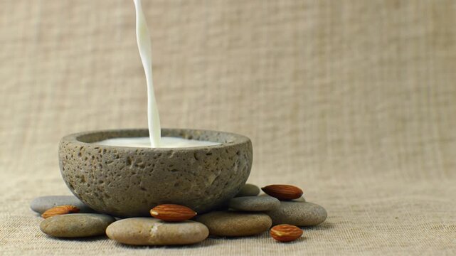 Almond milk pouring into stone bowl surrounded by almonds and pebbles on burlap background