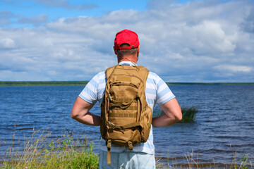 mature man with a backpack stands alone on the shore of the reservoir and enjoys the scenery, view from the back. summer walk, hiking and relaxation from urban conditions concept. © Dmitrii Masquerade