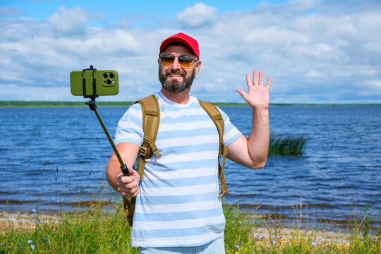 Travel blogger with backpack recording video on smartphone mounted on selfie stick while waving at camera on lakeshore. social media, travel content, adventure and tourism.