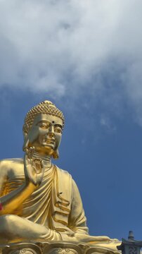 Vertical view shows golden Buddha statue with raised symbolic mudra gesture against blue sky. Mindfulness practice.