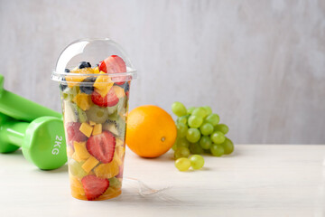 Fototapeta premium Tasty fruits in plastic cup, fork and dumbbells on wooden table, closeup. Space for text