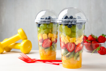 Fototapeta premium Tasty fruits in plastic cups, forks and dumbbells on light wooden table, closeup