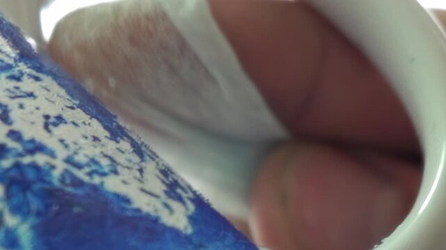 A close-up reveals a hand's delicate grip around a blue and white patterned ceramic mug. A soft white tissue cushions the finger as it gently cleans the rim in a morning moment.